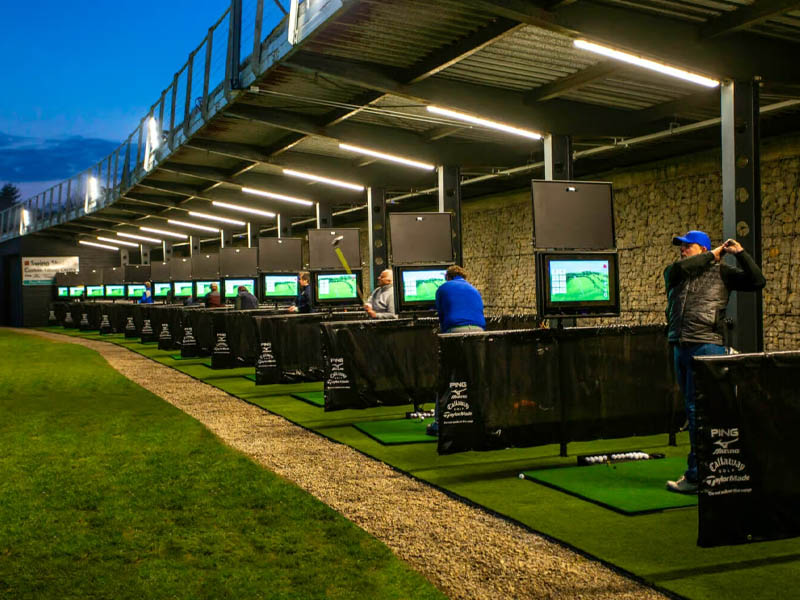 Golfers practicing in covered driving range bays equipped with TrackMan monitors and illuminated targets.