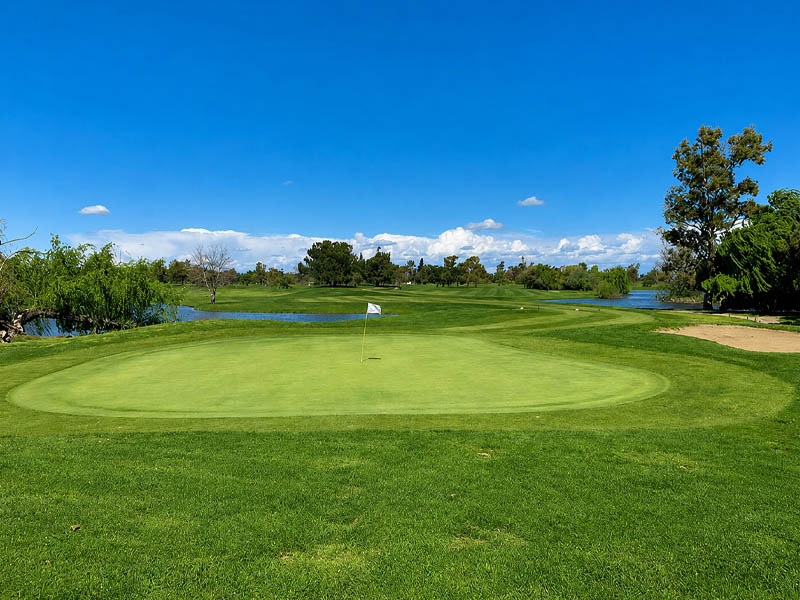 Putting green with a white flag, surrounding water hazards, and a sand bunker beneath a clear blue sky.