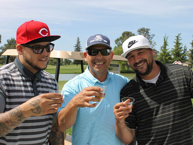 Three smiling golfers holding drinks together beside a golf cart during an outing at Micke Grove Golf Links.