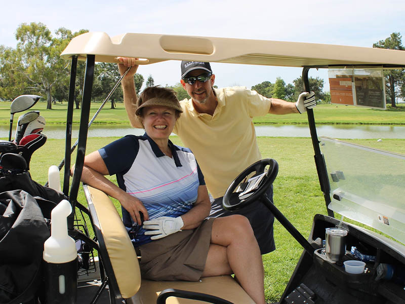 Two smiling golfers posing in and beside a golf cart near a pond at Micke Grove Golf Links.