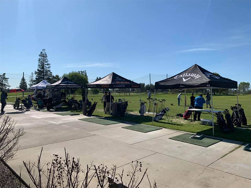 Golf equipment demonstration event with branded tents, club displays, practice mats, and golfers on the driving range.