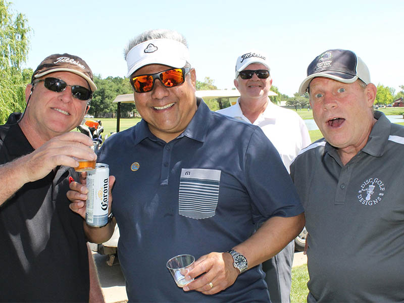 Four smiling golfers enjoying drinks together beside a golf cart during a sunny outing at Micke Grove Golf Links.