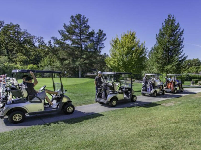 Group of golfers riding in golf carts along a paved path through a sunny, tree-lined golf course.