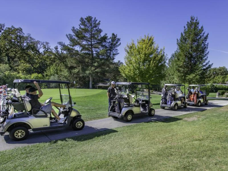 Group of golfers riding in golf carts along a paved path through a sunny, tree-lined golf course.
